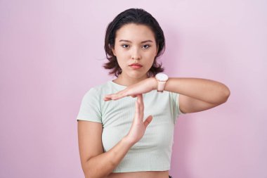 Pembe arka planda duran Latin kökenli genç bir kadın elleriyle jest yapıyor, hayal kırıklığına uğramış ve ciddi bir yüz ifadesi var. 