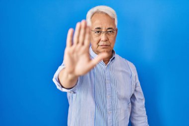 Hispanic senior man wearing glasses doing stop sing with palm of the hand. warning expression with negative and serious gesture on the face. 