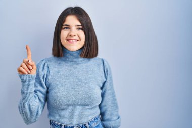 Mavi arka planda duran genç İspanyol kadın bir numarayı gösterip işaret ederken kendinden emin ve mutlu bir şekilde gülümsüyor.. 