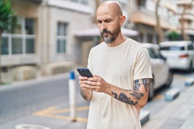 Genç kel adam akıllı telefon kullanıyor ve sokakta ciddi bir yüz ifadesi var.