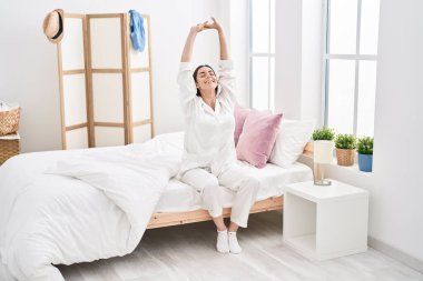 Young hispanic woman waking up sitting on bed at bedroom