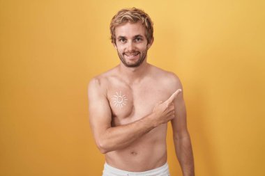 Beyaz adam üstsüz, neşeli bir güneş kremi sürmüş, yüzünde bir gülümseme, mutlu ve doğal bir ifadeyle elini ve parmağını yana kaldırmış. 