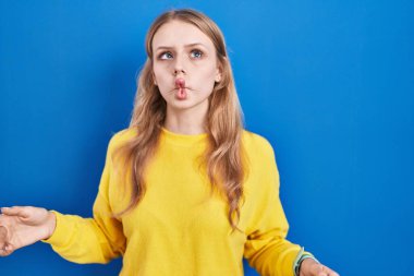 Young caucasian woman standing over blue background making fish face with lips, crazy and comical gesture. funny expression. 