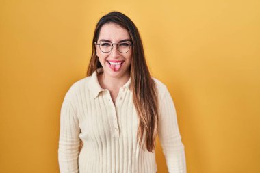Sarı arka planda dikilen genç İspanyol kadın komik bir ifadeyle mutlu bir şekilde dilini dışarı çıkarıyor. duygu kavramı. 