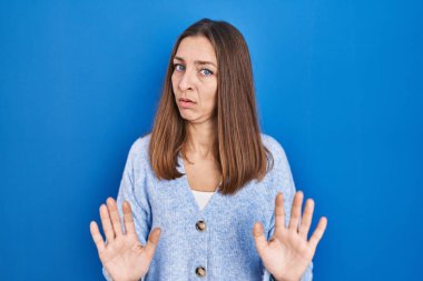 Young woman standing over blue background moving away hands palms showing refusal and denial with afraid and disgusting expression. stop and forbidden. 