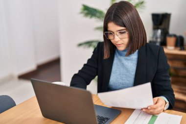 Young beautiful hispanic woman business worker using laptop reading document at office