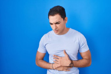 Mavi arka planda duran genç İspanyol adam midesi üstünde duruyor çünkü hazımsızlık, acı verici bir hastalık kendini iyi hissetmiyor. Ağrı kavramı. 