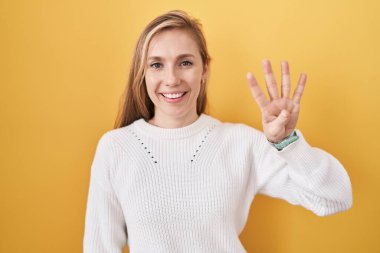 Sarı arka plan üzerine beyaz kazak giyen genç beyaz kadın dört numaralı parmaklarıyla gülümseyerek kendinden emin ve mutlu bir şekilde onu işaret ediyor.. 
