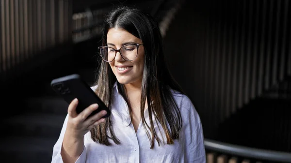 若い美しいヒスパニック女性はオフィスでスマートフォンを使用して幸せに微笑みます — ストック写真