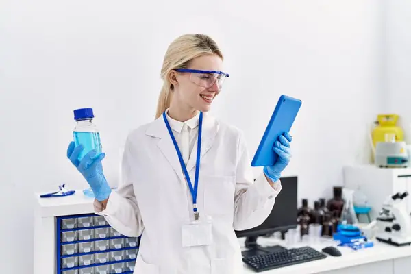 若い金髪女性科学者タッチパッドを使用して実験室で液体ボトルを保持 — ストック写真