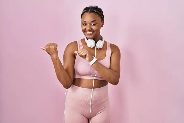 African american woman with braids wearing sportswear and headphones pointing to the back behind with hand and thumbs up, smiling confident