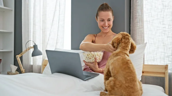 Young caucasian woman with dog watching movie on laptop sitting on bed at bedroom