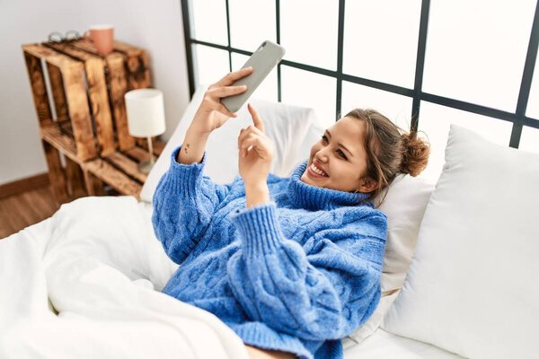 Young beautiful hispanic woman using smartphone lying on bed at bedroom