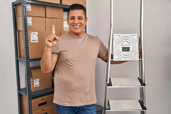 Hispanic young man working at small business ecommerce holding ladders smiling with an idea or question pointing finger with happy face, number one