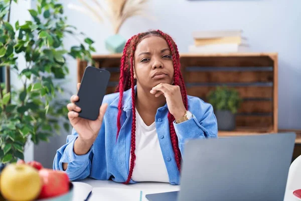 拿着辫子的非裔美国女人拿着智能手机 在屏幕上显示出一张空白的脸严肃地思考着手托着下巴的问题 思考着令人困惑的想法 — 图库照片