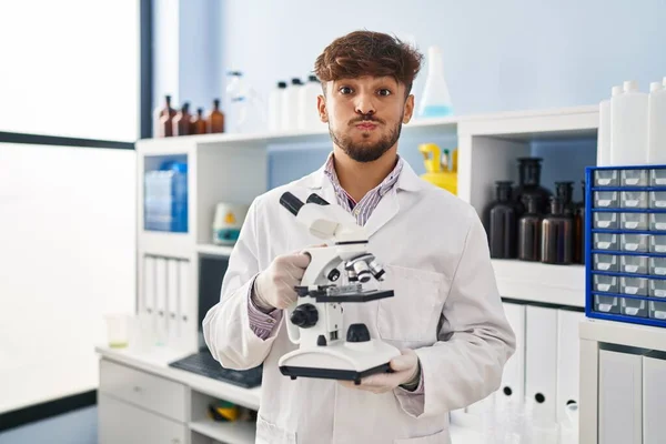 一名留着胡子的阿拉伯男子在科学家实验室工作 他拿着显微镜 用滑稽的脸鼓起双颊 嘴被空气吹胀 — 图库照片