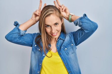 Mavi arka planda dikilen genç sarışın kadın komik ve çılgın poz veriyor parmakları tavşan kulağı gibi, neşeli gülümsüyor. 