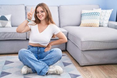 Oturma odasında yerde oturan beyaz bir kadın kameraya bakıp zafer işareti yaparken gülümsüyor. İki numara.. 