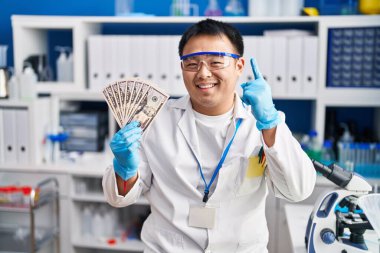 Laboratuvarda çalışan genç Çinli bir adam elinde para tutarken bir fikir veya soruyla gülümsüyor ve mutlu bir yüzle işaret ediyor. 