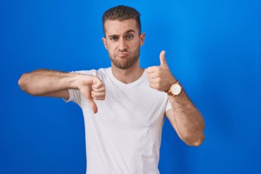 Kafkasyalı genç adam mavi arka planda duruyor. Başparmaklarını aşağı yukarı kaldırıyor. Anlaşmazlık ve uzlaşma ifadesi. çılgın çatışma 