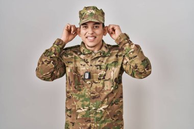 Genç Arap adam kamuflaj üniforması giyip gülümsüyor kulakları parmaklarıyla çekiyor, komik bir jest. deneme sorunu 