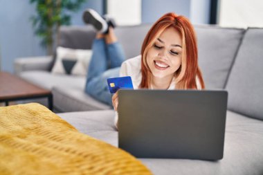 Evdeki kanepede dizüstü bilgisayar ve kredi kartı kullanan beyaz bir kadın.