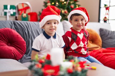 Evde Noel ağacının yanında kendinden emin bir şekilde oturan iki çocuk.