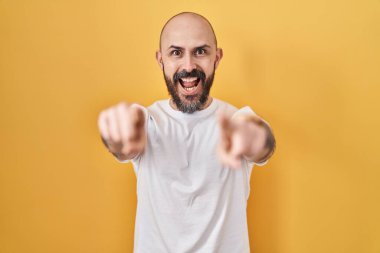 Sarı arka planda duran İspanyol dövmeli genç adam parmaklarıyla seni ve kamerayı işaret ediyor, neşeli ve pozitif gülümsüyor. 