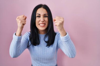 Pembe arka planda öfkeli ve kızgın duran İspanyol kadın öfkeyle bağırırken yumruklarını kaldırıyor ve sinirleniyor. öfke ve agresif konsept. 