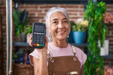 Çiçekçide çalışan, elinde veri bankası olan orta yaşlı bir kadın. Olumlu ve mutlu görünüyor. Kendine güvenen bir gülümsemeyle dişlerini gösteriyor. 