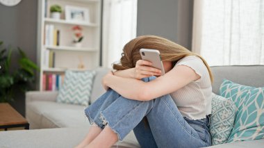 Akıllı telefon kullanan genç sarışın kadın evde stresli bir şekilde koltukta oturuyor.