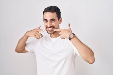 Beyaz arka planda durup gülümseyen, parmaklarıyla ve ağzıyla işaret eden yakışıklı İspanyol. diş sağlığı kavramı. 