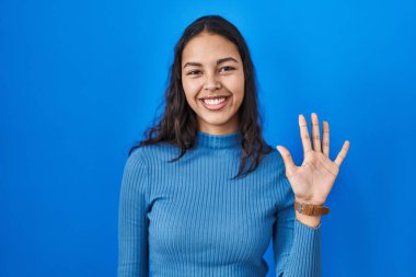 Mavi arka planda duran Brezilyalı genç kadın beş numarayı gösterip işaret ederken kendinden emin ve mutlu bir şekilde gülümsüyor.. 