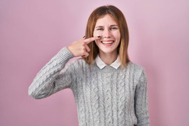 Pembe arka planda duran güzel bir kadın. Parmakları burnuna ve yüzüne dönük, neşeli bir şekilde gülümsüyor. güzellik konsepti 