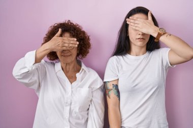İspanyol anne ve kızı pembe arka plan üzerine gündelik beyaz t-shirt giyiyorlar. Gözleri elle kaplıyorlar, ciddi ve üzgün görünüyorlar. Görme, saklama ve reddetme konsepti 