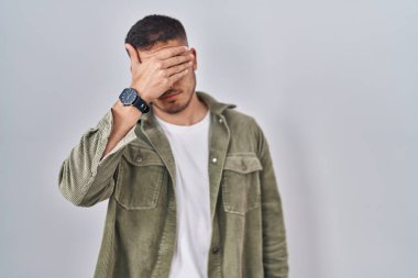 Young hispanic man standing over isolated background covering eyes with hand, looking serious and sad. sightless, hiding and rejection concept 