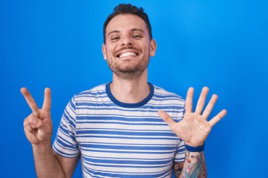Young hispanic man standing over blue background showing and pointing up with fingers number seven while smiling confident and happy. 