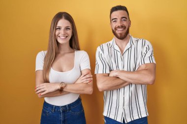 Sarı arka planda duran genç çift kollarını kavuşturup kameraya bakarken gülümsüyor. pozitif kişi. 