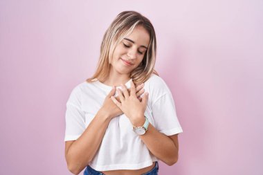 Pembe arka planda duran genç sarışın kadın elleri göğsünde, gözleri kapalı ve yüzünde minnet dolu bir jest ile gülümsüyor. sağlık kavramı. 
