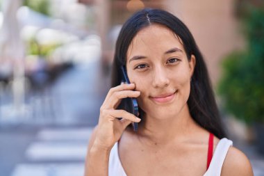 Genç, güzel, İspanyol bir kadın gülümsüyor ve caddede akıllı telefondan konuşuyor.