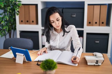 Young caucasian woman business worker writing on notebook using touchpad and earphones at office