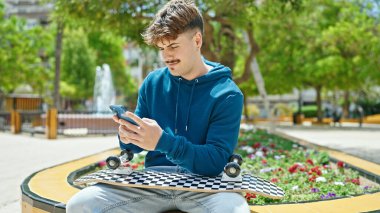 Parkta akıllı telefon kullanarak kaykay yapan genç bir İspanyol.