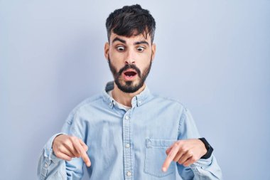 Mavi arka planda duran sakallı genç İspanyol adam parmaklarını göstererek reklamını yapıyor, şaşırmış yüzünü ve ağzını açıyor. 