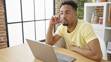Afro-Amerikalı adam laptopunu masaya oturmuş yemek odasında düşünüyor.