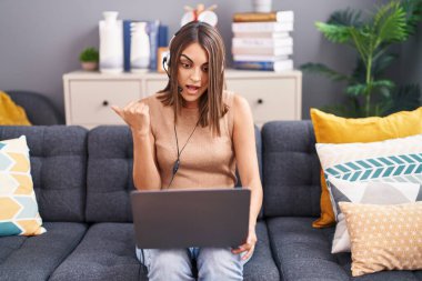 Evde çalışan genç İspanyol kadın operatör kulaklığı takıyor başparmağıyla yan tarafa bakıyor ve ağzı açık gülümsüyor. 