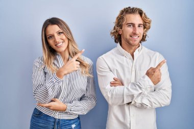 Mavi arka planda duran genç çift, yüzünde kocaman bir gülümsemeyle, kameraya el ile işaret ederek bakıyor.. 