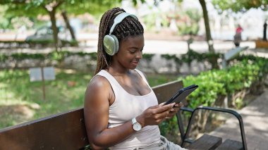 Parktaki bankta oturan dokunmatik ped ve kulaklık kullanan Afrikalı Amerikalı kadın.