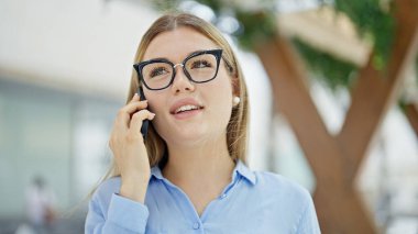 Genç, sarışın, iş kadını, gülümseyen, kendine güvenen, akıllı telefondan konuşan biri.