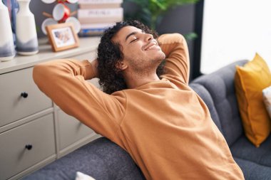 Young latin man relaxed with hands on head sitting on sofa at home