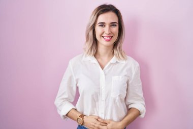 Young beautiful woman standing over pink background with hands together and crossed fingers smiling relaxed and cheerful. success and optimistic 